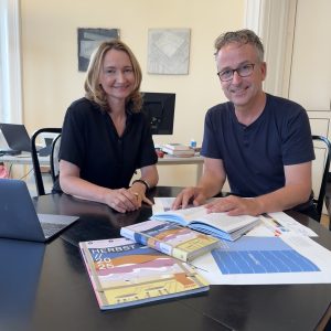 Elisabeth Stein-Hölzl und Matthias Opis hinter einem Tisch mit der Verlagsvorschau und einem Buch. Sie lächeln in die Kamera.