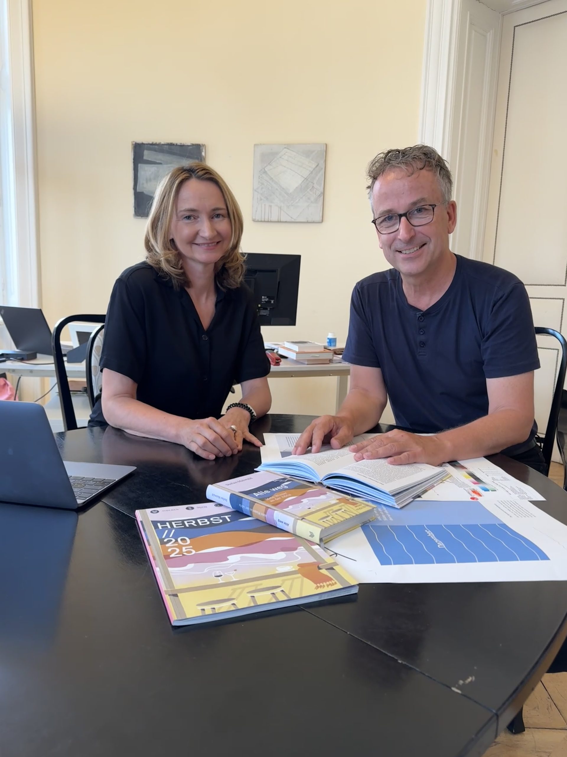 Elisabeth Stein-Hölzl und Matthias Opis hinter einem Tisch mit der Verlagsvorschau und einem Buch. Sie lächeln in die Kamera.
